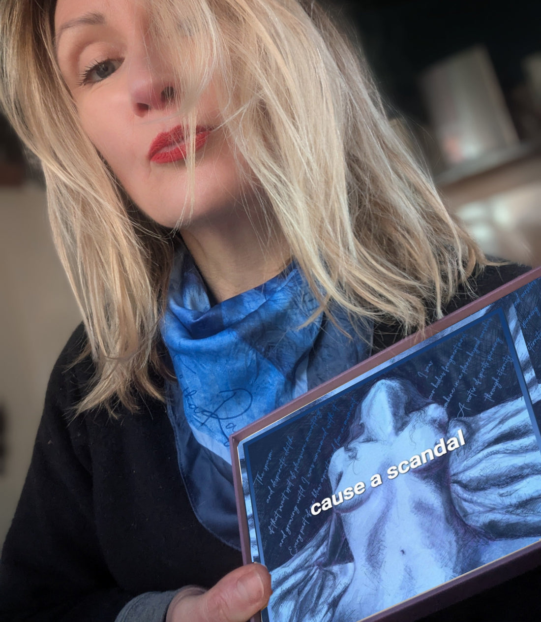 Tabitha Rayne a white woman with blond hair and red lipstick holds up a box with a blue silk scarf featuring a nude silk scark - it says cause a scandal on it. 